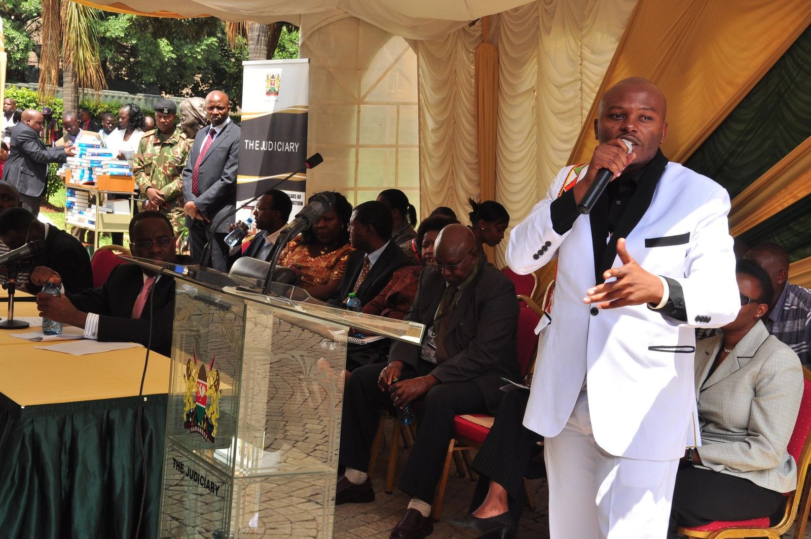 Jay MauMau performing at a Judiciary of Kenya event, in a white suit with a microphone